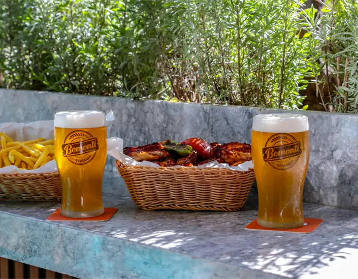 Two cold beers on square drink coasters beside a basket of fries and grilled food on an outdoor patio table.