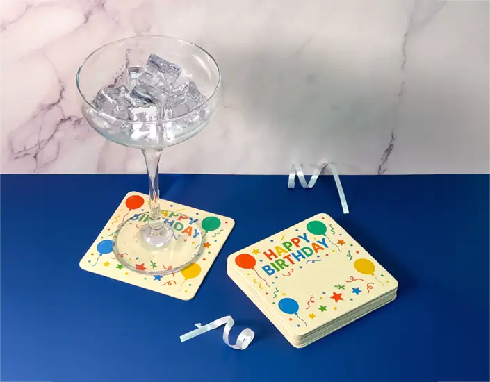 Happy Birthday drink coasters stacked beside a cocktail glass, styled as party favors for an adult birthday celebration.