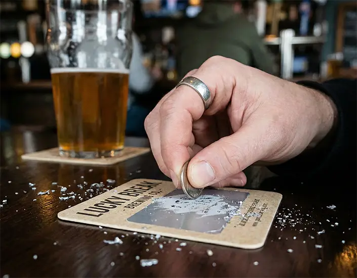Customer scratching a scratch-and-win drink coaster on a bar table beside a glass of beer, revealing a prize message during an in-venue promotion.