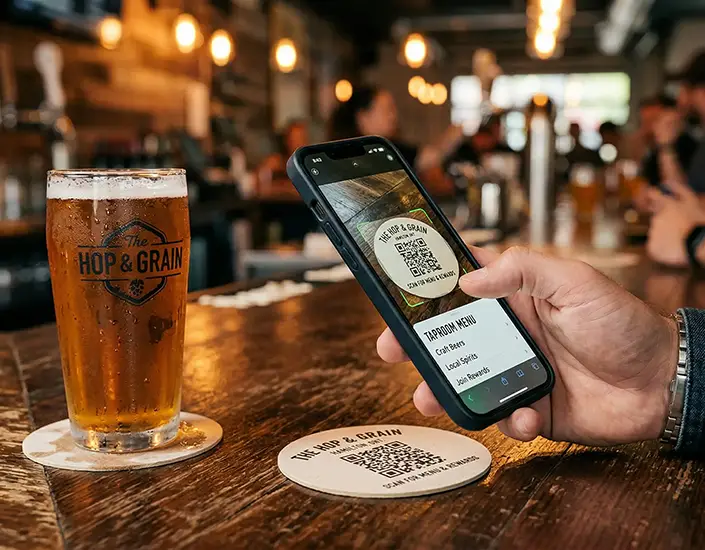 Customer scanning QR code on branded bar coaster next to beer at busy taproom