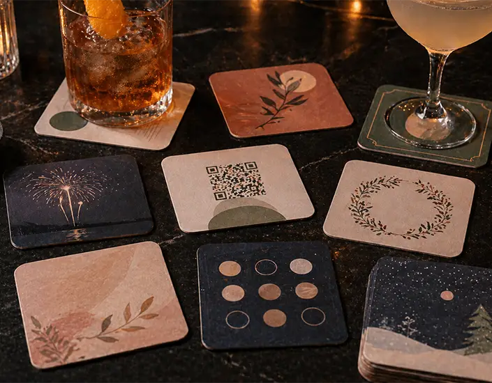Custom pulpboard coasters with various designs arranged on a bar table, showing promotional styles including QR code.