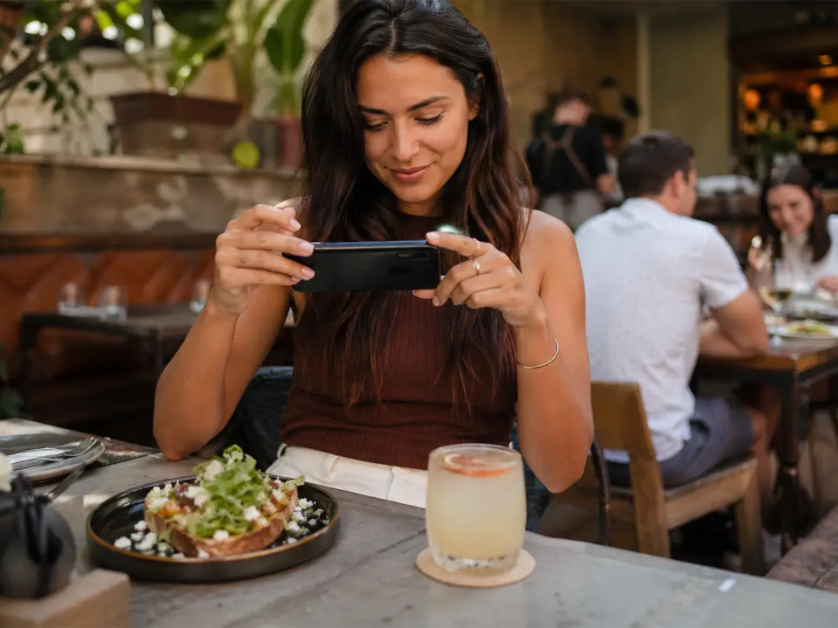 Food influencer photographing a meal and cocktail at a restaurant table with a branded coaster under the drink.