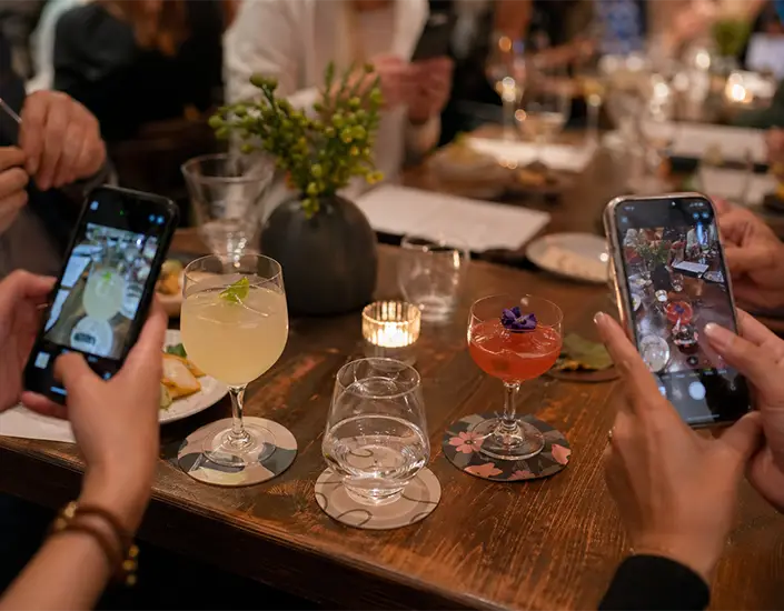 Diners taking photos of cocktails on branded coasters during a restaurant gathering with multiple drinks on the table.