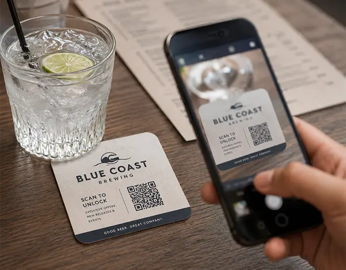 Blue Coast Brewing coaster with a QR code being scanned by a smartphone, showing how coasters connect offline marketing with digital engagement.