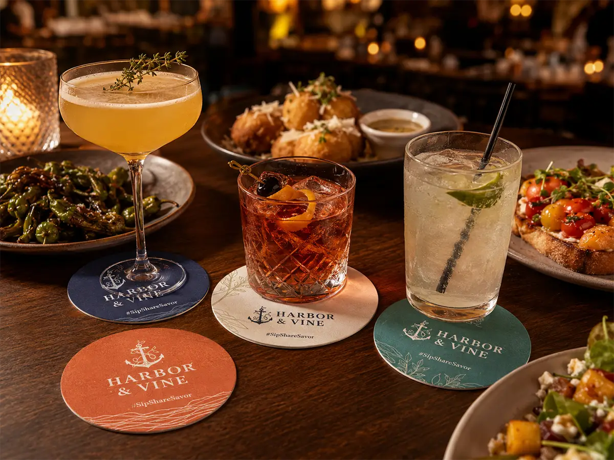 Restaurant table with drinks, food, and custom Harbor & Vine coasters featuring branded social media hashtag designs.