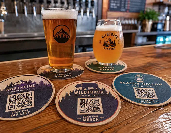 Custom brewery QR code coasters displayed on a bar with beer glasses and scan prompts for tap list, events, and merch.