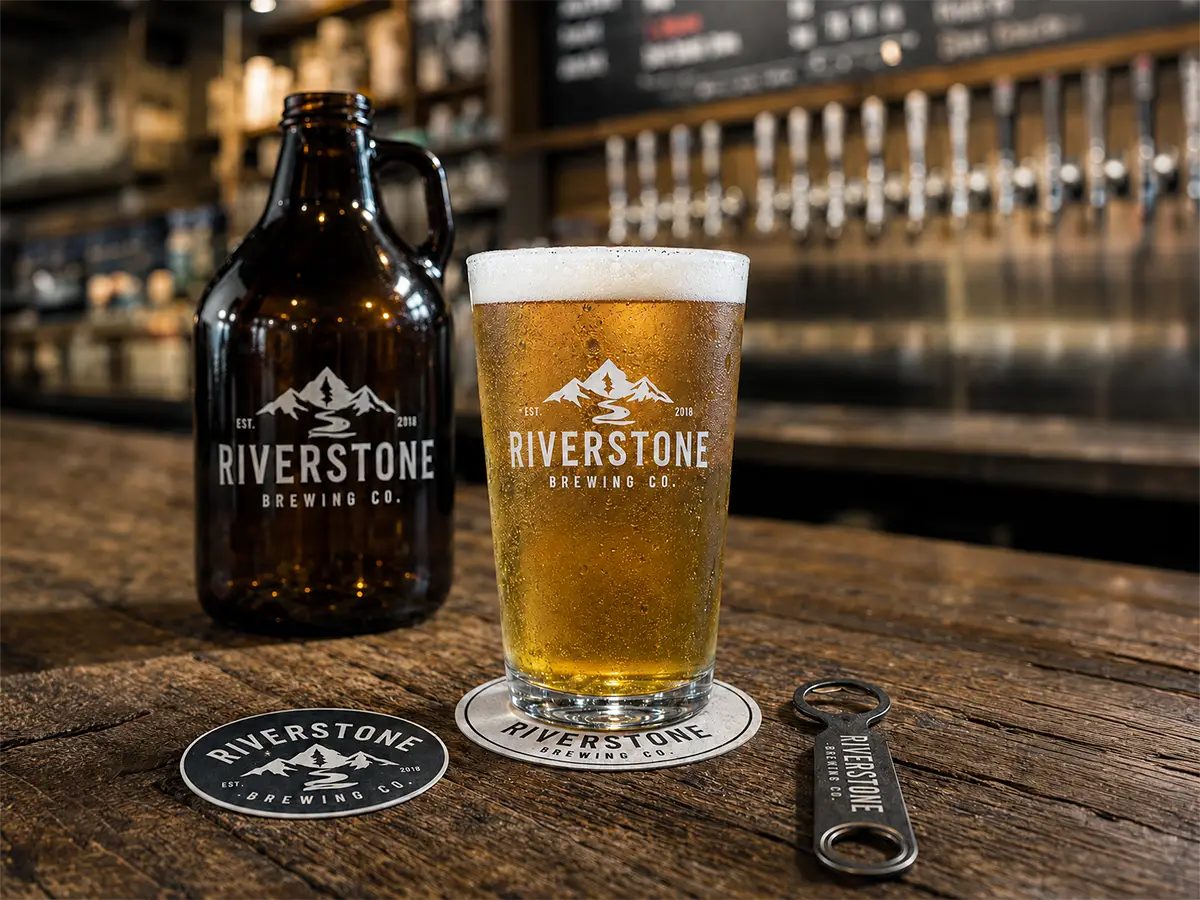Custom brewery promotional products including a branded beer glass, growler, coasters, and bottle opener displayed on a rustic taproom table.