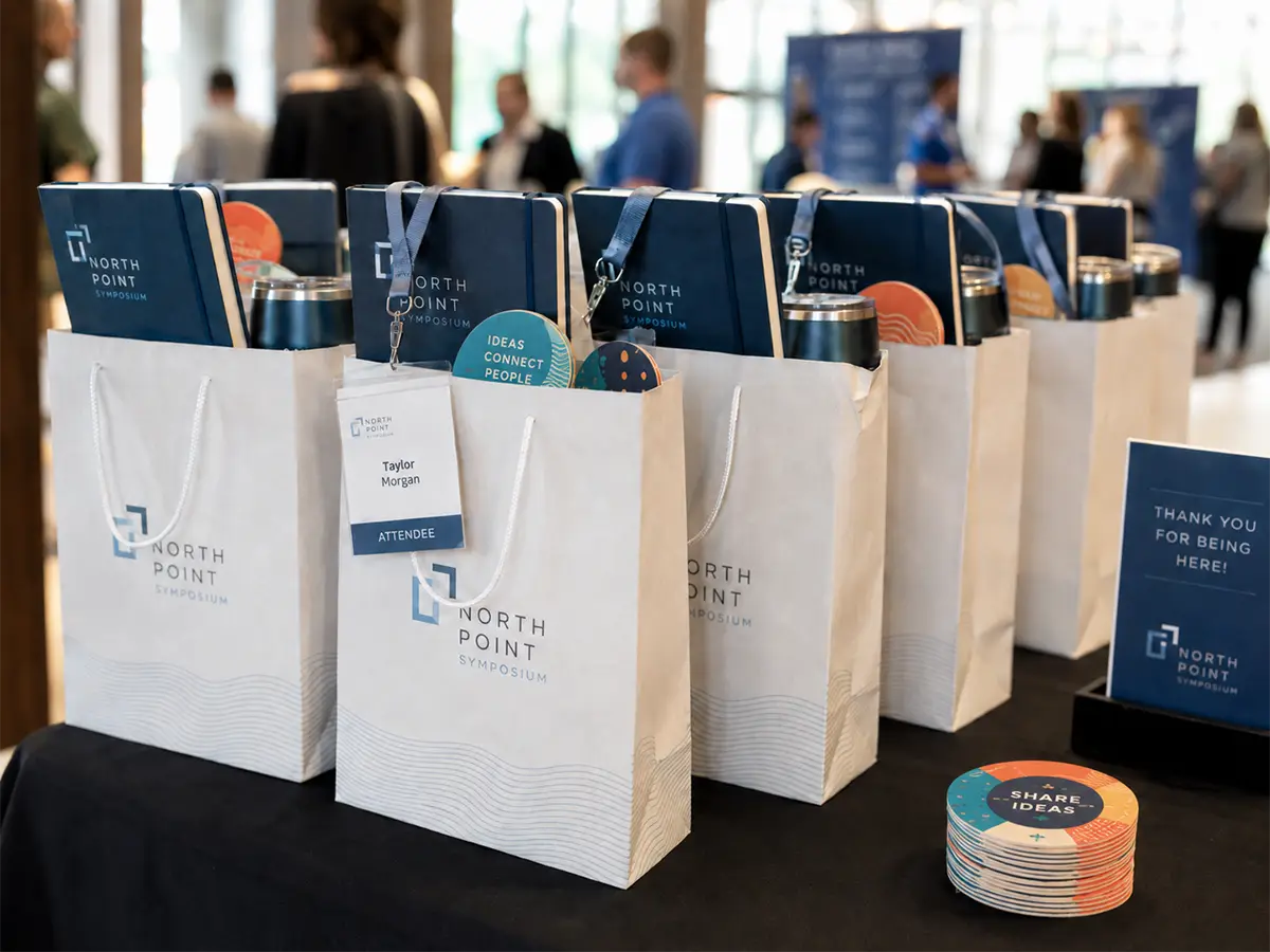 Conference swag bags lined up on a welcome table with notebooks, drinkware, badges, and custom coasters.