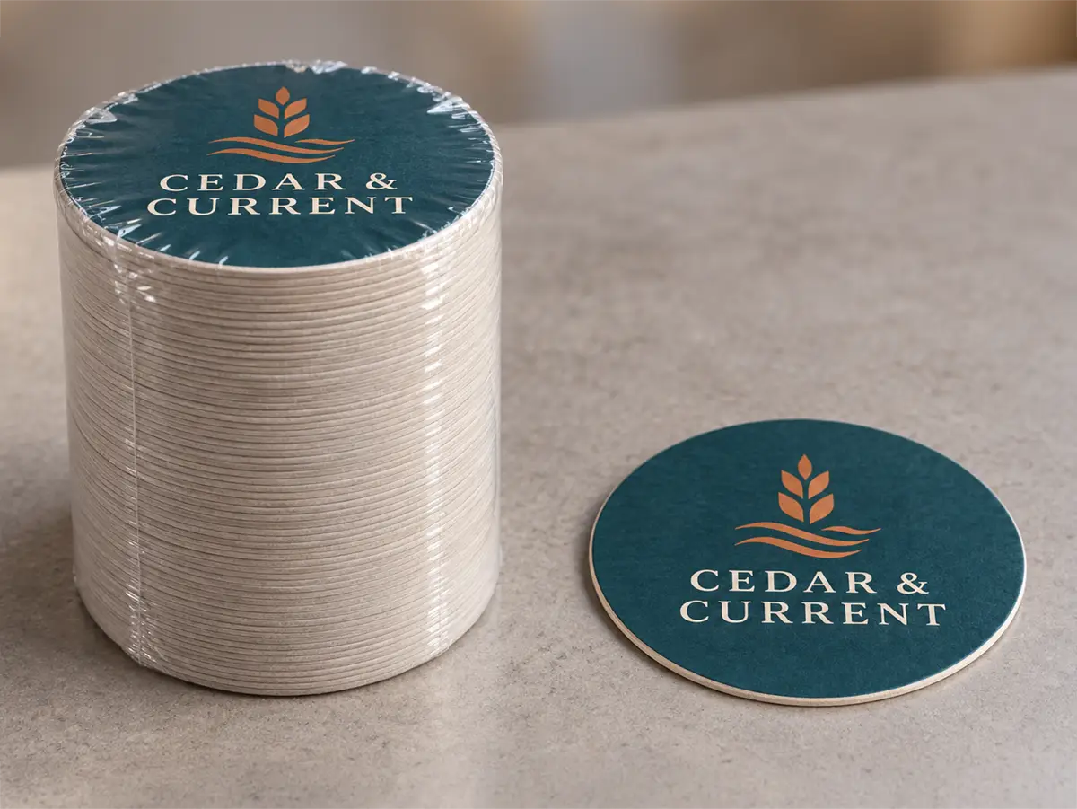 Shrink-wrapped stack of round Cedar & Current pulpboard coasters beside a matching loose coaster.